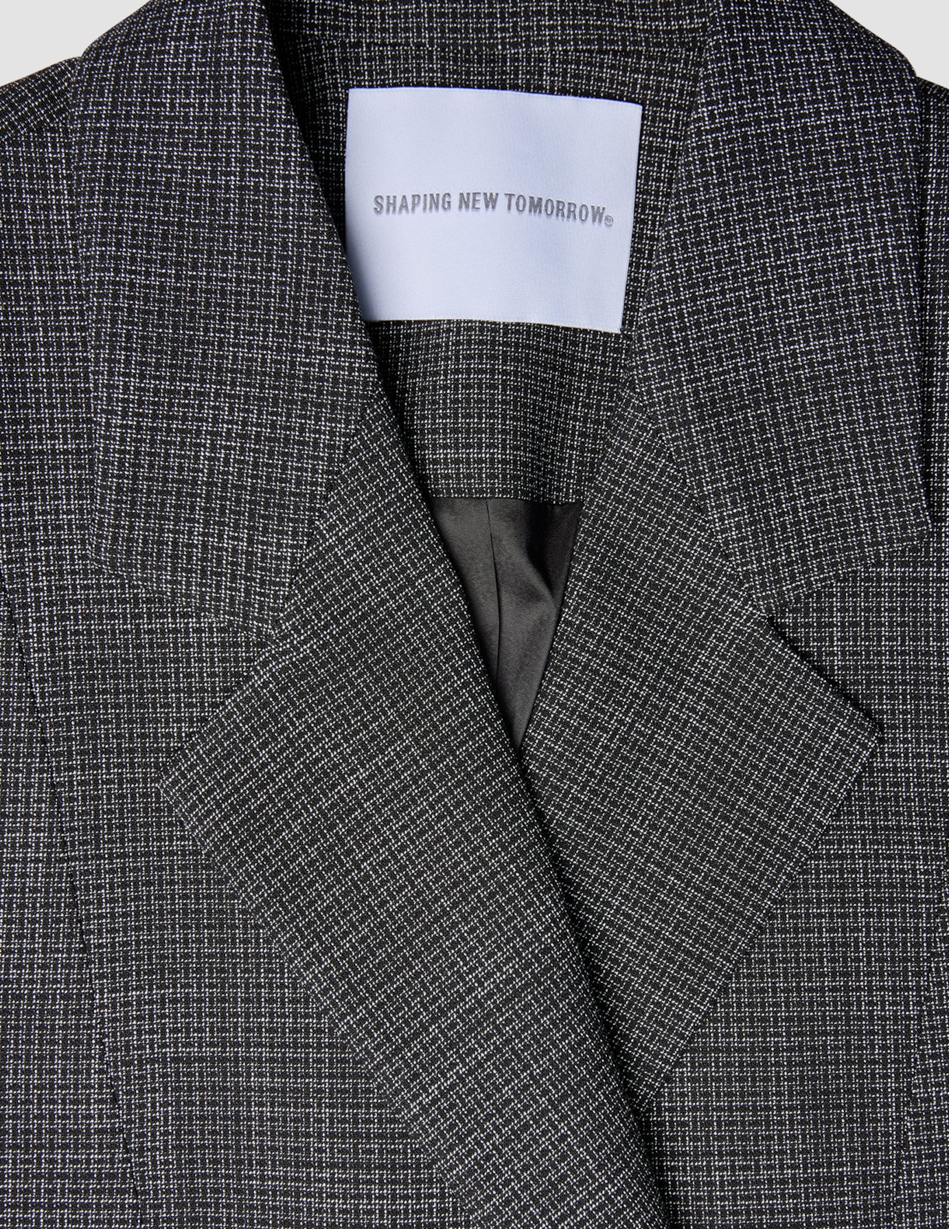 Nærbillede af en sort-hvid ternet Women Nara Blazer Fitted Black Check med et SHAPING NEW TOMORROW-mærke ved halsudskæringen.